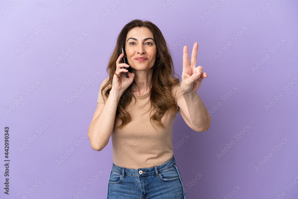Middle aged caucasian woman using mobile phone isolated on purple background smiling and showing victory sign