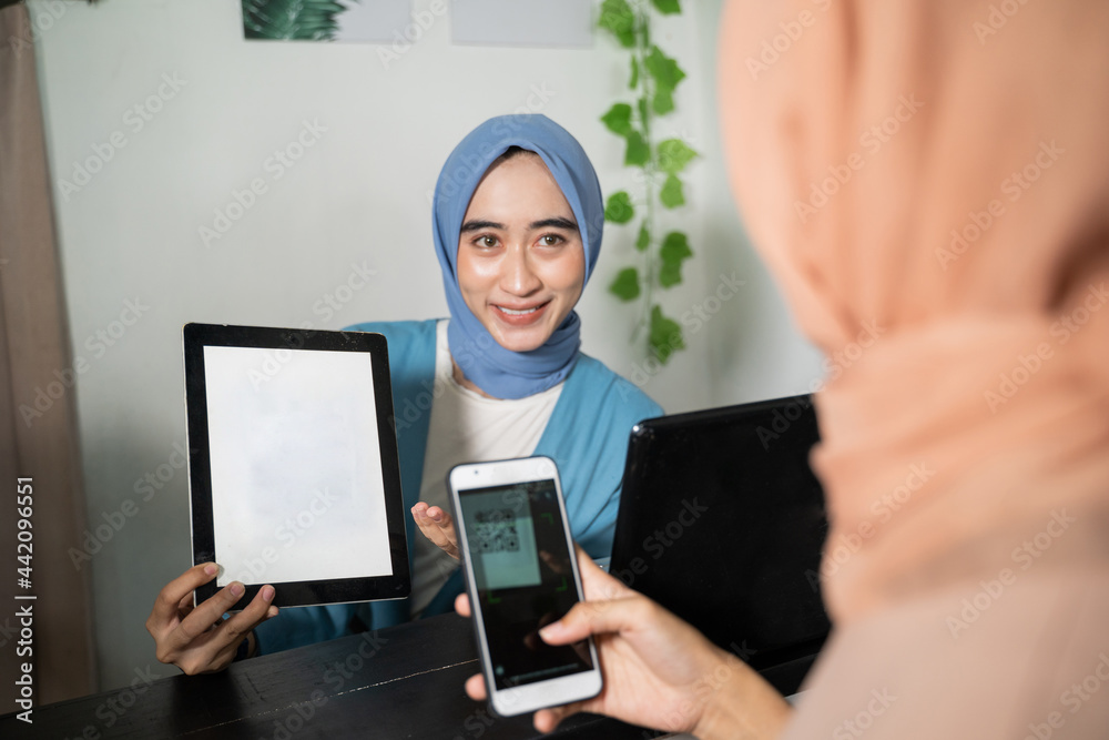 A hijab business woman holds a tablet and shows the screen to customers who use cellphones while paying non-cash by scanning barcodes at the cashier at a boutique shop