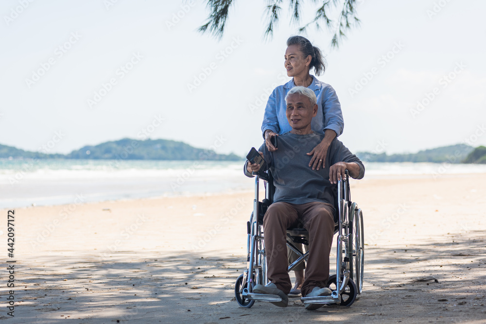 Happy Asian senior man on wheelchair and elderly woman couple smiling on sand of the beach for taking the patient travel together