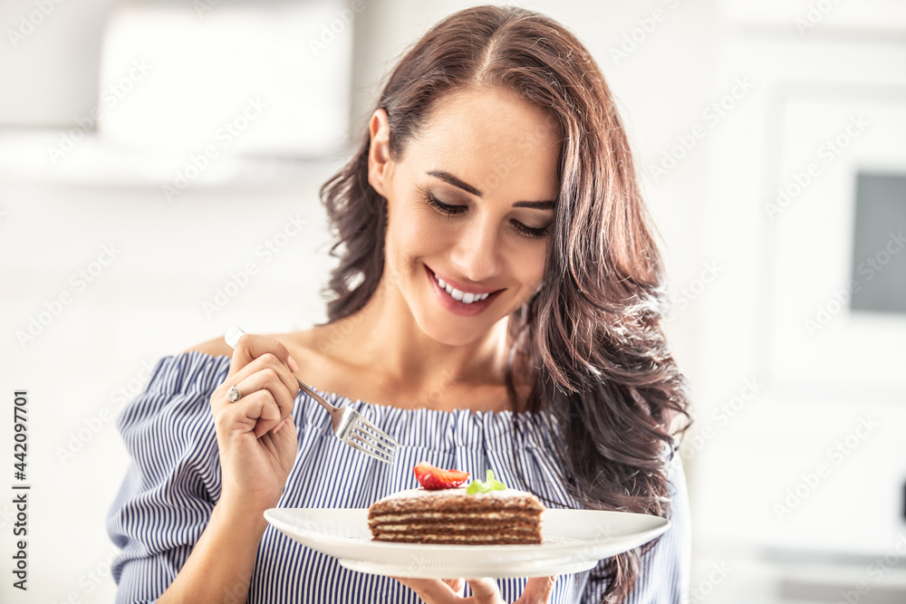Layered brown adn white cake with strawberry and mint leaf on top held by a woman with a fork Layered brown adn white cake with strawberry and mint leaf on top held by a woman with a fork