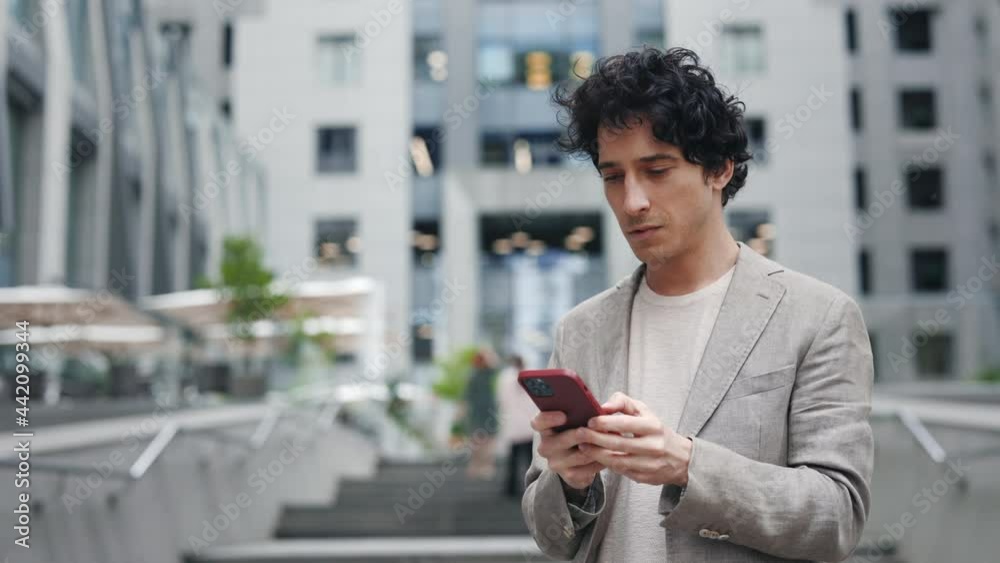 Success. Move camera view of the businessman standing in the city center street and using his phone while texting, scrolling and tapping with smile. Technology and communication concept. Success. Move camera view of the businessman standing in the city center street and using his phone while texting, scrolling and tapping with smile. Technology and communication concept.