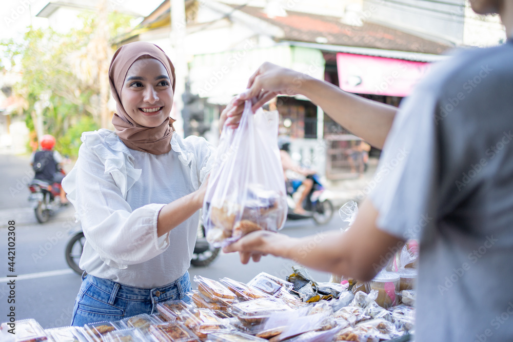 A beautiful girl in a headscarf takes a takjil food order with a plastic bag from a roadside stall seller