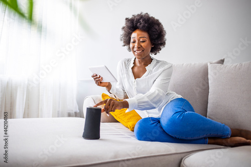 Black woman connecting phone to her virtual assistant smart speaker. Happy black woman using smart speaker at home. Young cheerful woman controlling home devices with a voice commands