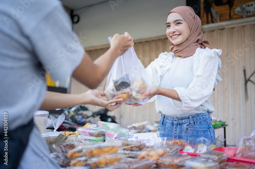 A beautiful girl in a headscarf gives a takjil food order in a plastic bag to a buyer at a roadside stall