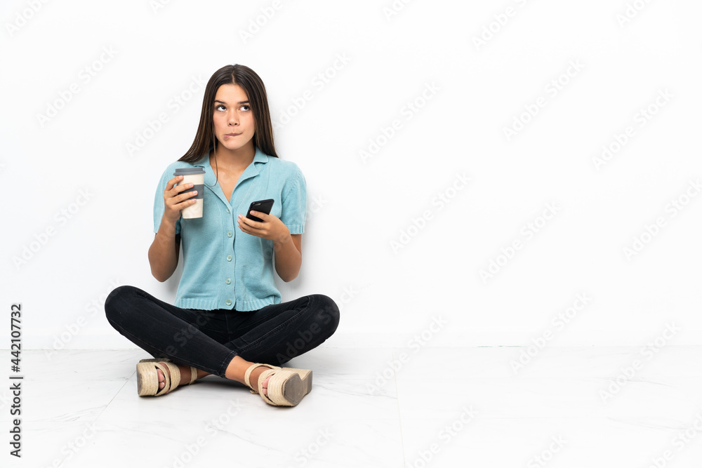 Teenager girl sitting on the floor holding coffee to take away and a mobile while thinking something Teenager girl sitting on the floor holding coffee to take away and a mobile while thinking something