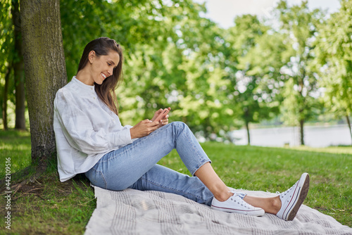 Happy young woman using mobile phone in the park sitting on the blanket