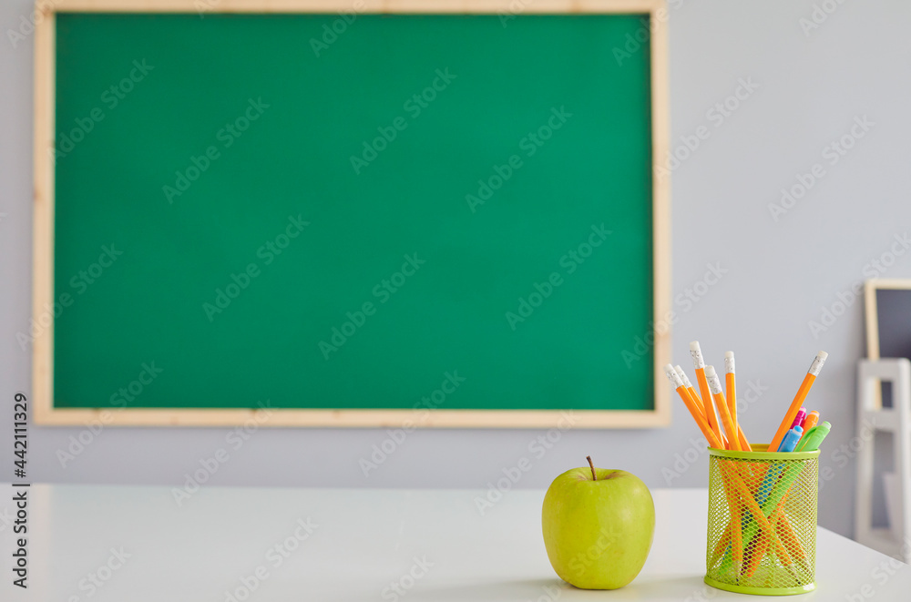 Green apple and stationery on teacher's desk at classroom, empty space for your text on chalkboard. Fruit and school supplies on table
