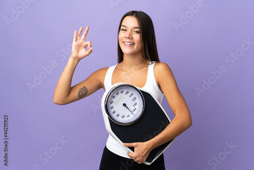 Teenager girl isolated on purple background holding a weighing machine and doing OK sign