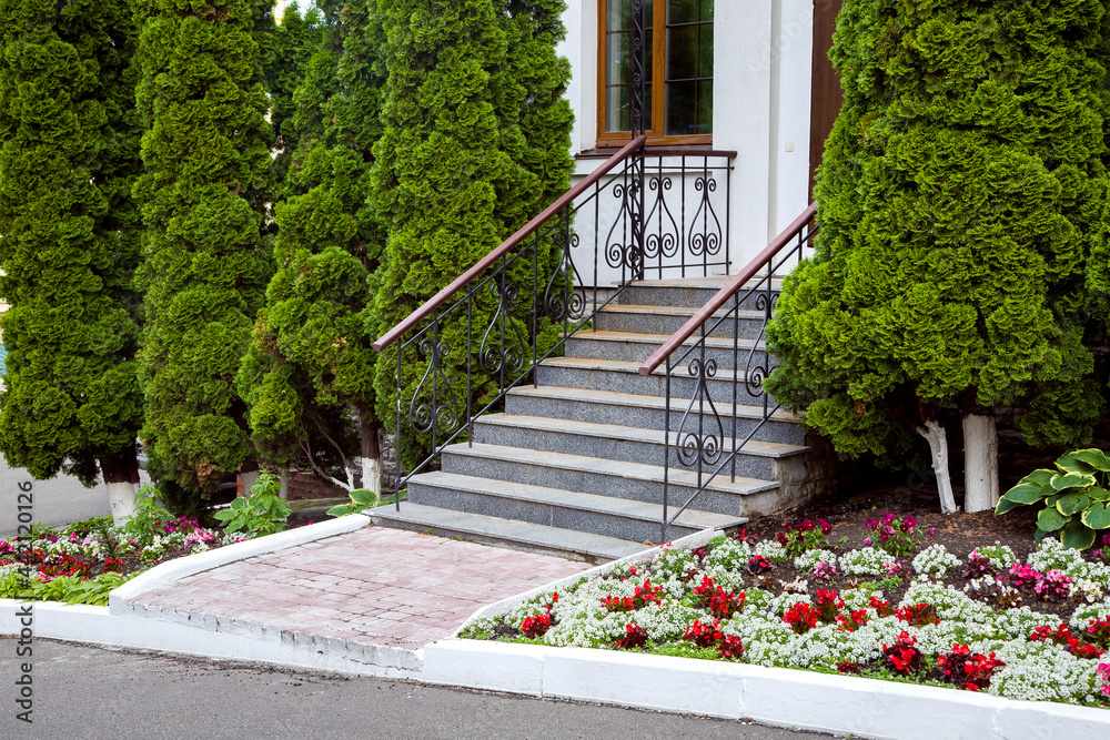 a granite staircase with a wrought iron railings, entrance steps, exit to the backyard with a flowerbed with flowers and evergreen thuja trees, nobody.
