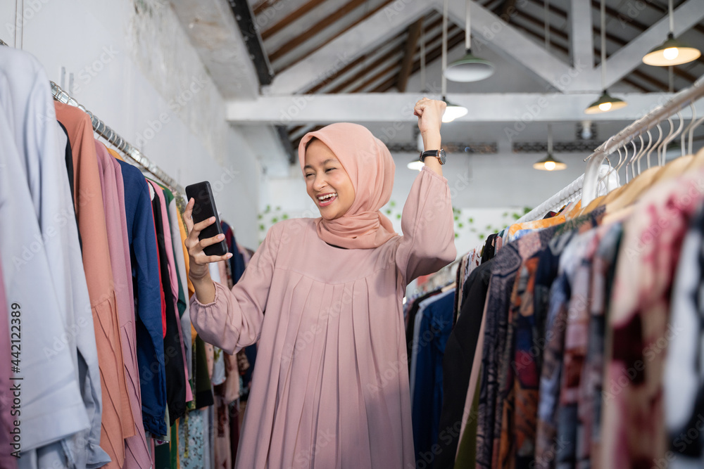 a beautiful girl in a headscarf felt excited to raise her hand while holding a smartphone while standing in a boutique shop