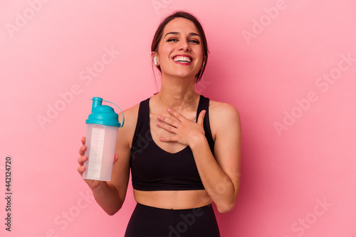 Young caucasian woman drinking a protein shake isolated on pink background laughs out loudly keeping hand on chest.