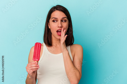 Young caucasian woman holding an ice cream isolated on blue background is saying a secret hot braking news and looking aside