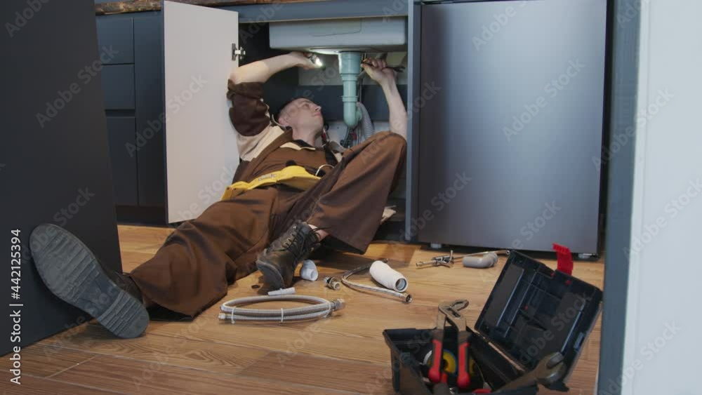 Handheld tracking shot with slowmo of male plumber in uniform lying under kitchen sink and fixing leak or installing new pipes
