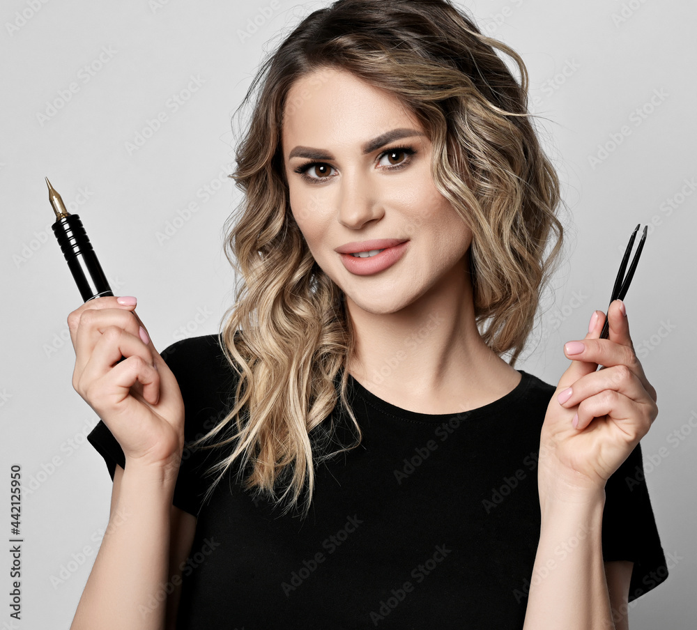 Obraz premium Portrait of smiling young curly blonde woman in black t-shirt with copy space, holding permanent makeup machine in hand and tweezers for eyebrows tattooing over light background