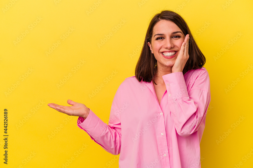Young caucasian woman isolated on yellow background holds copy space on a palm, keep hand over cheek. Amazed and delighted. Young caucasian woman isolated on yellow background holds copy space on a palm, keep hand over cheek. Amazed and delighted.