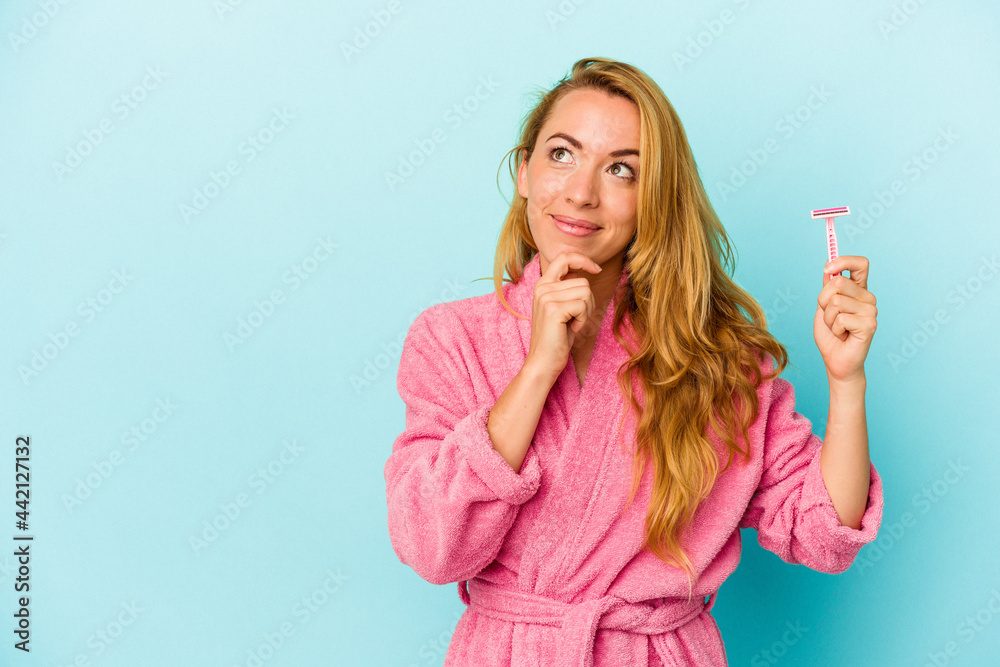 Caucasian woman wearing a bathrobe holding razor blade isolated on pink background looking sideways with doubtful and skeptical expression. Caucasian woman wearing a bathrobe holding razor blade isolated on pink background looking sideways with doubtful and skeptical expression.