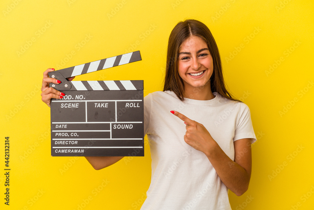 Young caucasian woman holding clapperboard isolated on white background smiling and pointing aside, showing something at blank space.