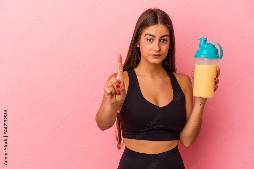 Young caucasian woman holding a protein shake isolated on yellow background showing number one with finger. Young caucasian woman holding a protein shake isolated on yellow background showing number one with finger.