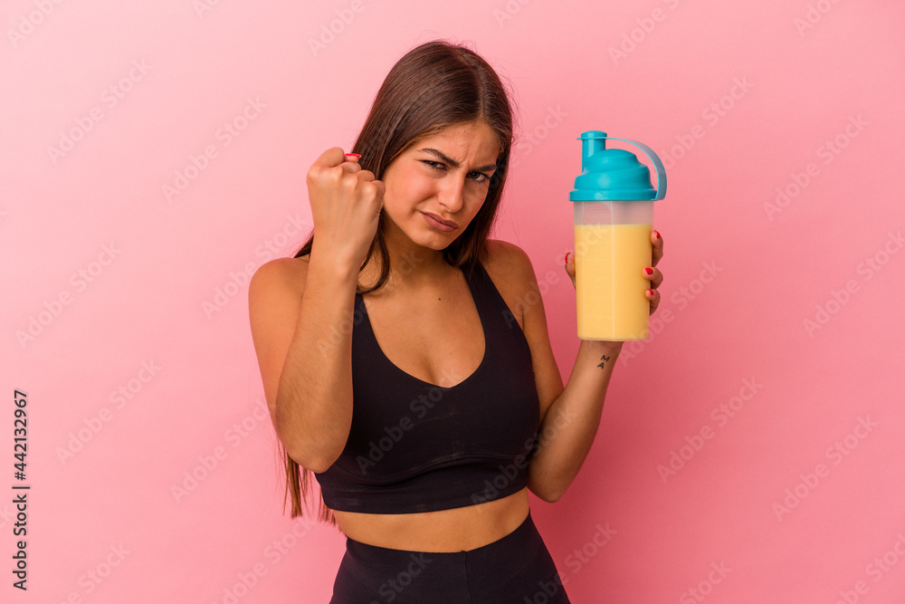 Young caucasian woman holding a protein shake isolated on yellow background showing fist to camera, aggressive facial expression.