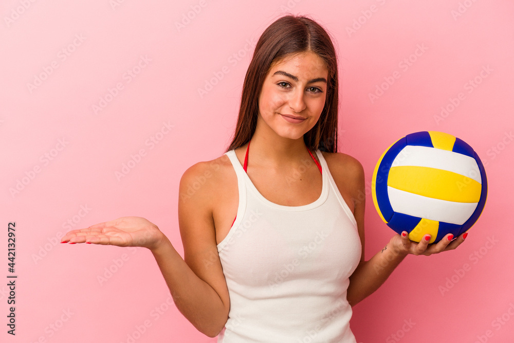 Young caucasian woman holding a volleyball ball isolated on pink background showing a copy space on a palm and holding another hand on waist.