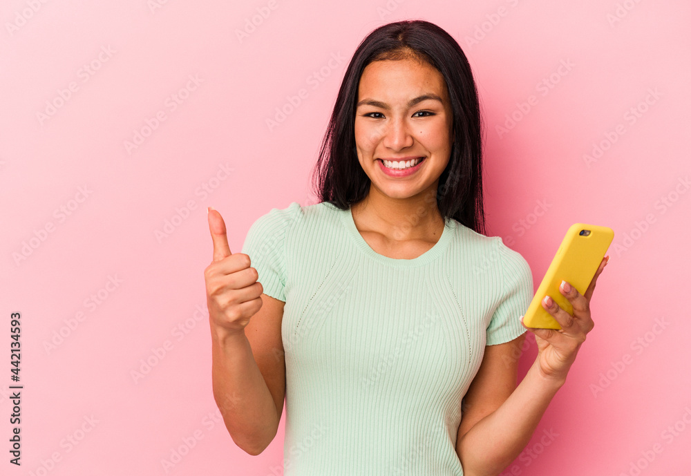 Young Venezuelan woman holding a mobile phone isolated on pink background smiling and raising thumb up Young Venezuelan woman holding a mobile phone isolated on pink background smiling and raising thumb up
