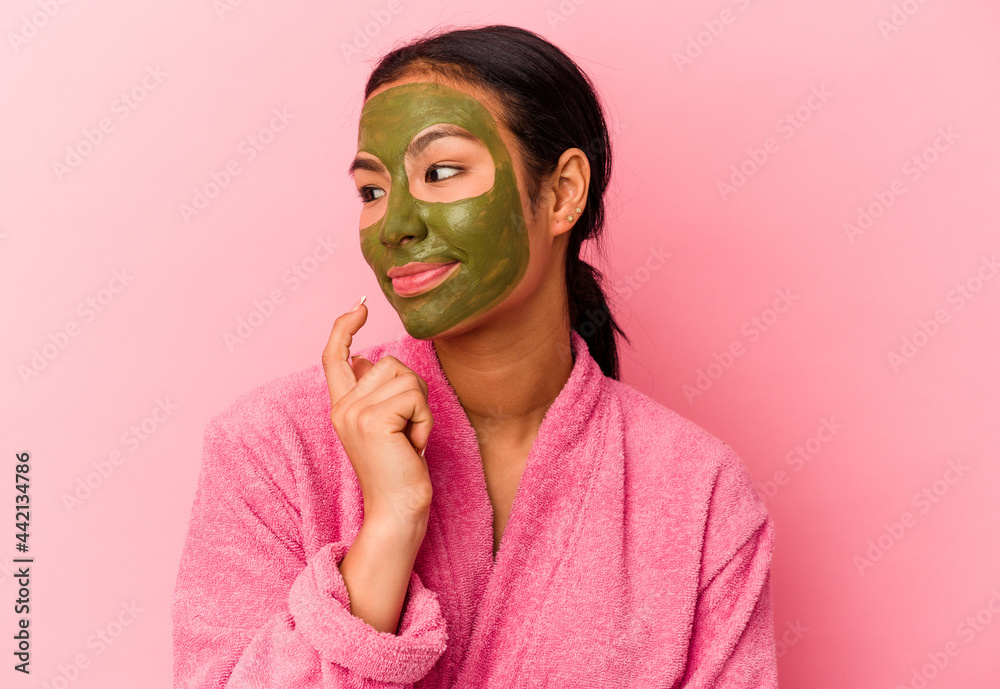 Young Venezuelan woman wearing a bathrobe and facial mask isolated on pink background relaxed thinking about something looking at a copy space. Young Venezuelan woman wearing a bathrobe and facial mask isolated on pink background relaxed thinking about something looking at a copy space.