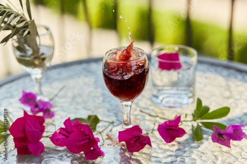 Explosion of elegant and colorful glass of red wine, wine splash in the glass. Summer cocktails on the background. Decorative pink flowers and a branch of an olive tree in a glass. 