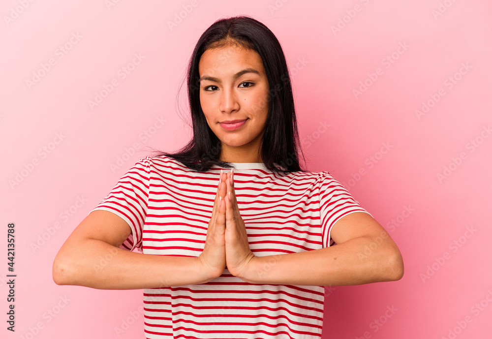 Young Venezuelan woman isolated on pink background praying, showing devotion, religious person looking for divine inspiration.