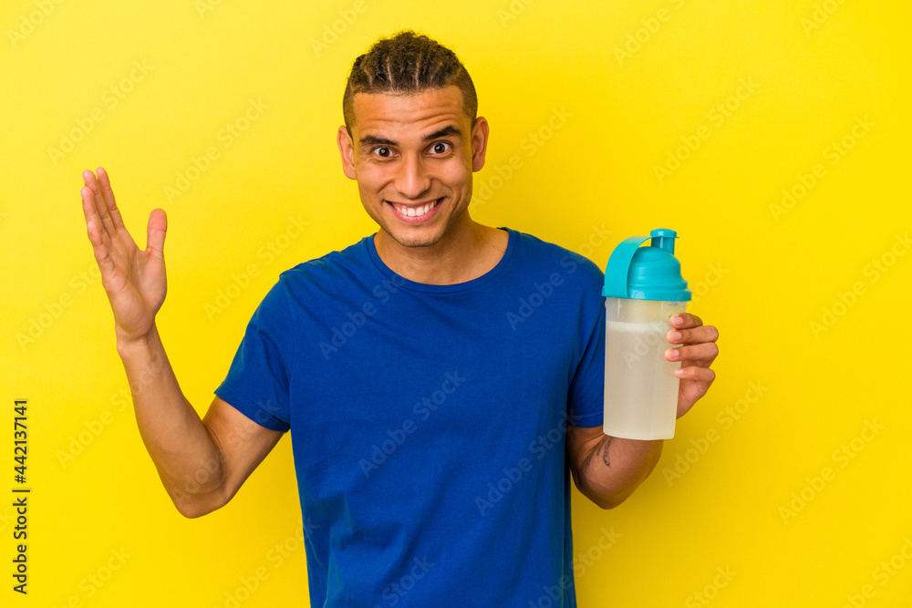 Young venezuelan man drinking a protein shake isolated on yellow background receiving a pleasant surprise, excited and raising hands. Young venezuelan man drinking a protein shake isolated on yellow background receiving a pleasant surprise, excited and raising hands.
