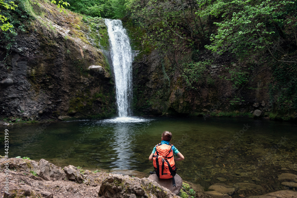 Obraz premium Child boy schoolboy with hiking backpack sitting alone in forest near lake and watching waterfall. Children's exploration and curiosity of nature, adventure learning and outdoor education