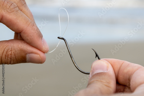 Hands of a white man running a nylon fishing line into the eye of a black fishhook in front of the sea. Mention to coastal fishing, sport and leisure with the family on a sunny weekend.