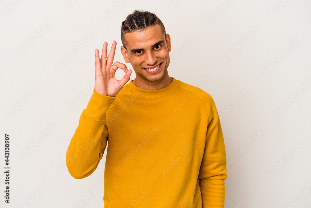 Young venezuelan man isolated on white background winks an eye and holds an okay gesture with hand. Young venezuelan man isolated on white background winks an eye and holds an okay gesture with hand.