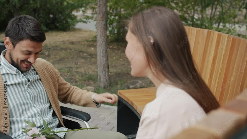 Disabled man moving wheelchair to young woman giving flower holding hands talking laughing enjoying romantic date outside. Relationship and youth concept. Disabled man moving wheelchair to young woman giving flower holding hands talking laughing enjoying romantic date outside. Relationship and youth concept.