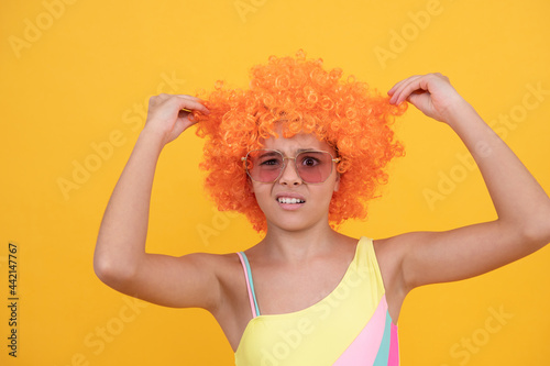 unhappy child in sunglasses and swimsuit does not like orange wig on yellow background, dislike