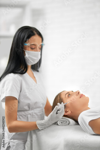 Side view of young brunette beautician in protective mask doing special beauty injection for young attractive woman. Concept of process tighten skin from wrinkles in professional beautician.