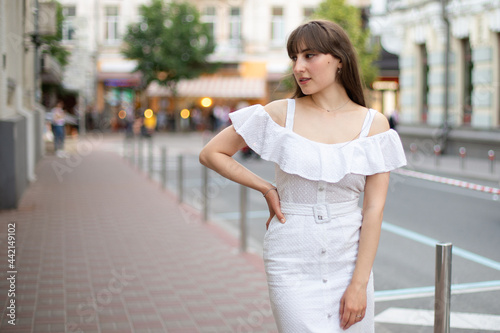 close-up of a beautiful woman with brown hair in a white dress who looks to the side on a summer street background. place for your design