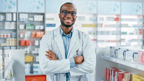 Pharmacy: Professional Confident Black Pharmacist Wearing Lab Coat and Glasses, Crosses Arms and Looks at Camera Smiling Charmingly. Druggist in Drugstore Store with Shelves Health Care Products