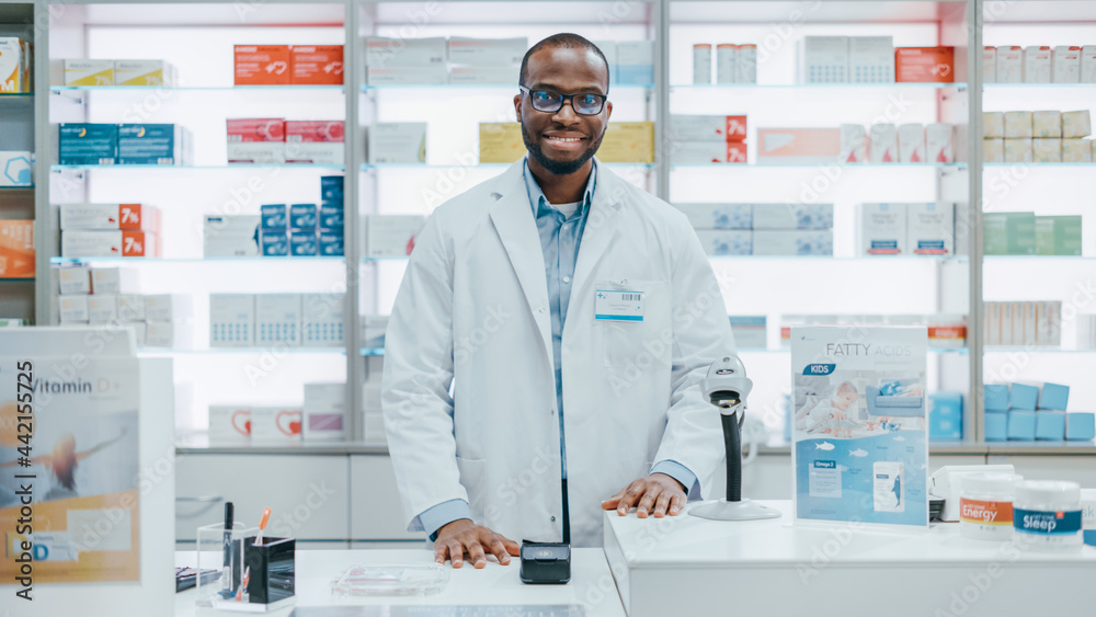 Pharmacy Drugstore Checkout Counter: Portrait of Handsome Professional Black Male Pharmacist Wearing White Lab Coat, Looks at Camera, Smiles. Shelves with Medicine Packages, Health Care Products