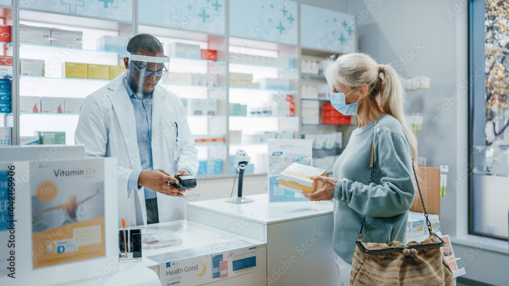 Obraz premium Pharmacy Drugstore Checkout Counter: Professional Black Pharmacist Wearing Face Shield Sells Medicine to Senior Female Customer, who is wearing Face Mask, Use Contactless Payment. Coronavirus Safety