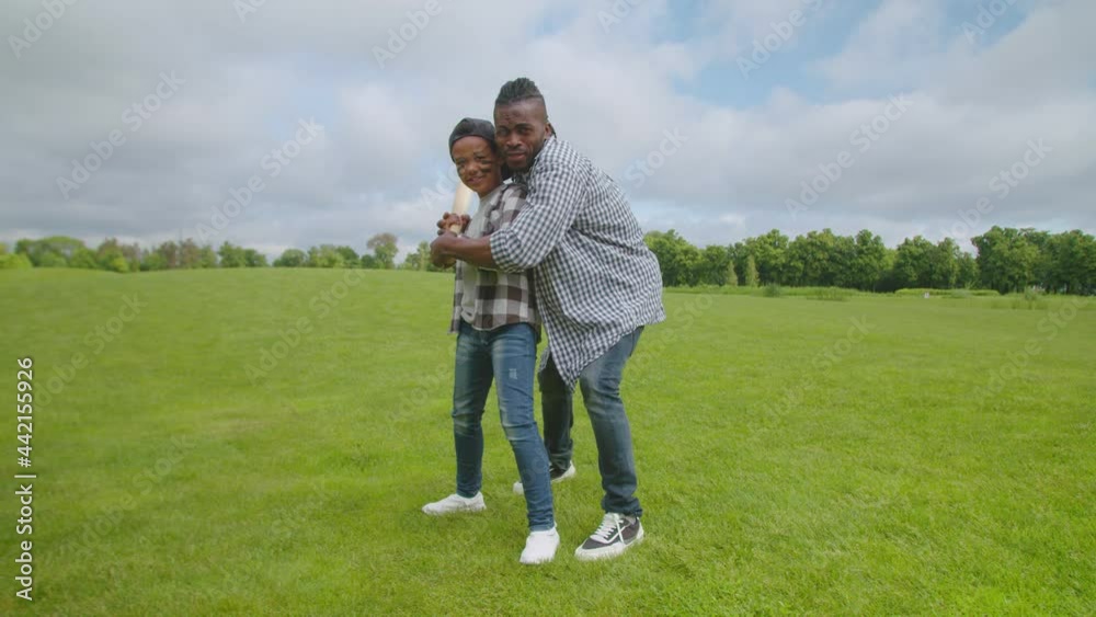 Affectionate handsome african american father teaching cute preadolescent son bat swing, showing right grip of baseball bat while enjoying leisure together and playing baseball in public park. Affectionate handsome african american father teaching cute preadolescent son bat swing, showing right grip of baseball bat while enjoying leisure together and playing baseball in public park.