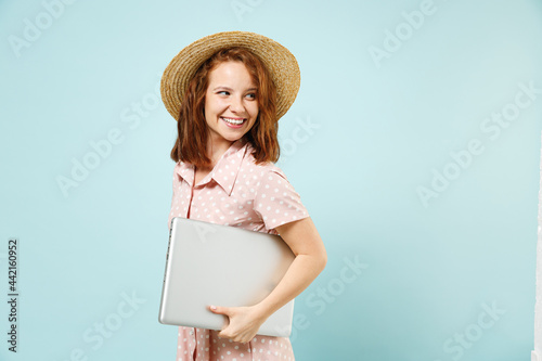 Side view smiling happy young redhead curly woman 20s wears casual pink dress straw hat hold laptop pc computer under her hand look saide back isolated on pastel blue color background studio portrait. photo
