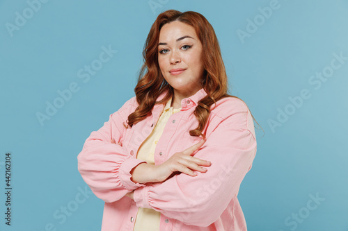 Young confident redhead chubby overweight woman 30s with curly hair in pink shirt casual clothes hold hand crossed folded isolated on pastel blue background studio portrait. People lifestyle concept photo