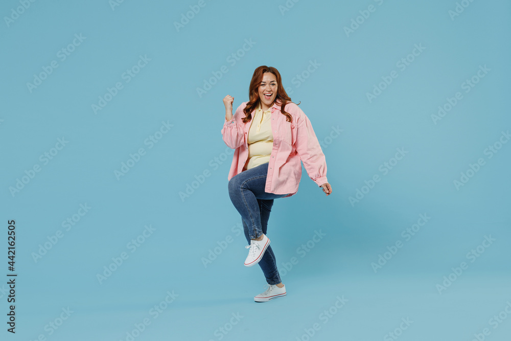 Full length young smiling redhead chubby overweight woman 30s with curly hair wear in pink shirt jeans casual clothes do winner gesture clench fist isolated on pastel blue background studio portrait Full length young smiling redhead chubby overweight woman 30s with curly hair wear in pink shirt jeans casual clothes do winner gesture clench fist isolated on pastel blue background studio portrait