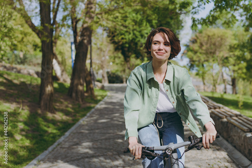 Young pensive dreamful happy woman 20s wearing casual green jacket jeans riding bicycle bike on sidewalk in city spring park outdoors, look aside People active urban healthy lifestyle cycling concept