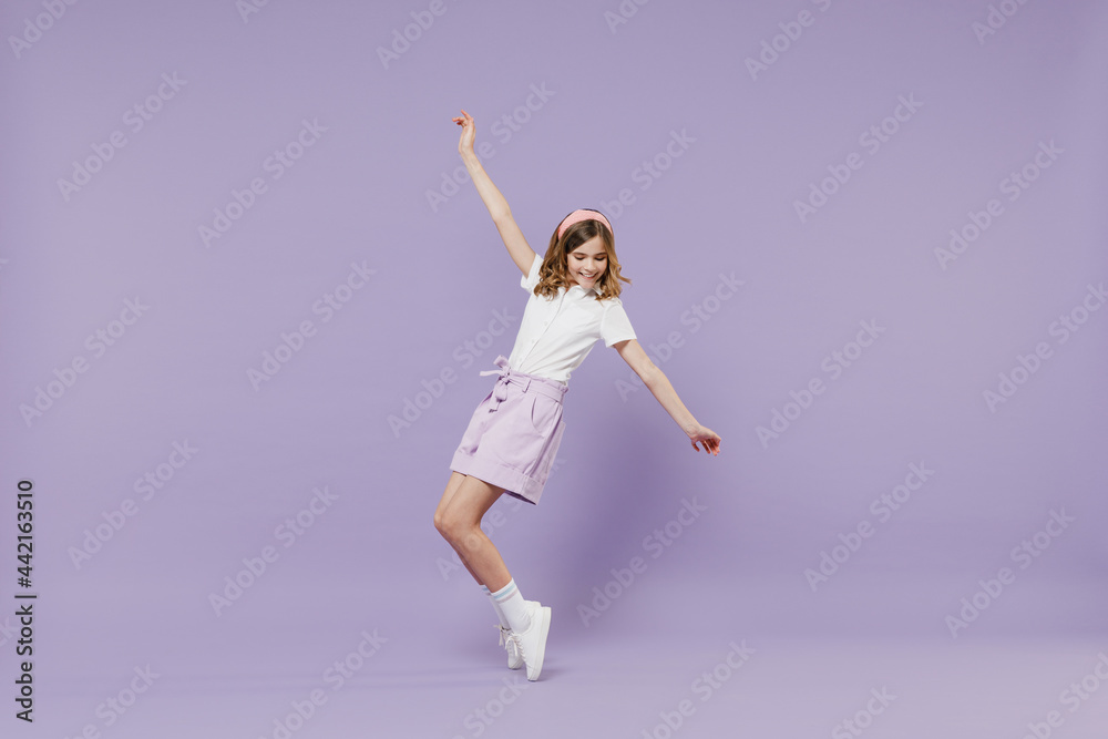 Full length little hppy blonde kid girl 12-13 year old in white shirt stand on toes with outstretched hands leaning back dance isolated on purple color background Childhood children lifestyle concept