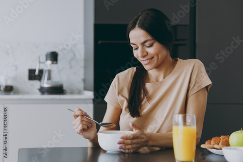 Photography Young calm smiling happy brunette housewife caucasian woman 20s wear casual clothes beige shirt eat oatmeal porridge muesli cooking food in light kitchen at home alone