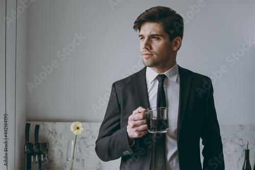 Young serious minded successful employee business man corporate lawyer 20s wearing formal black suit shirt tie drink coffee breakfast look aside in light kitchen Achievement career lifestyle concept.