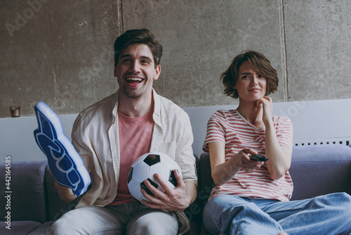 Couple friends sad woman happy man football fans 20s in casual clothes foam glove finger up support favorite team scream with soccer ball indoors at home flat watch tv People sport family concept.