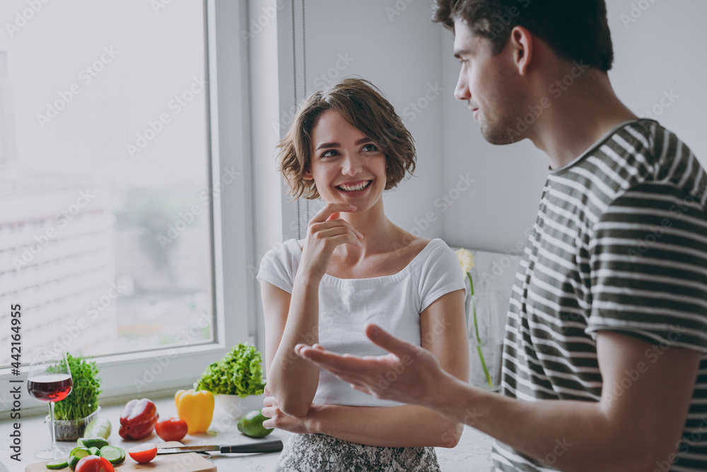 Young caucasian happy cheerful couple two woman man 20s wearing casual t-shirt clothes speak to each other cooking food in light kitchen at home together. Healthy diet lifestyle relationship concept.