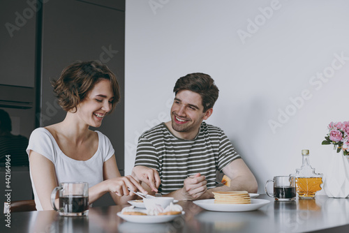 Young satisfied happy couple two caucasian woman man in casual t-shirt clothes sit by table eat pancakes with maple syrup cook food in light kitchen at home together Healthy diet lifestyle concept